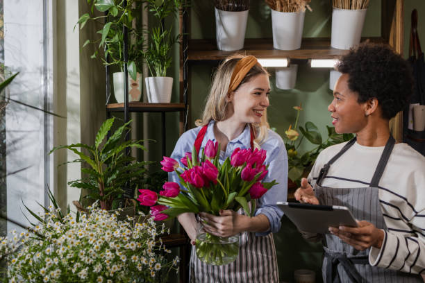 Portrait of two multiracial flower store workers looking at each other. A young blond woman is holding a vase with roses while a Latin American woman is holding a digital tablet.